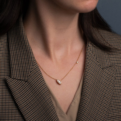 A close-up of a model wearing a delicate gold necklace with a marquise and round white lab-grown diamond pendants, styled with a brown and black houndstooth blazer.