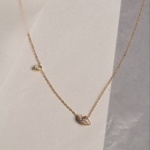 A delicate gold chain necklace with two pear-cut lab grown diamonds of different sizes, displayed on a textured white and gray surface.